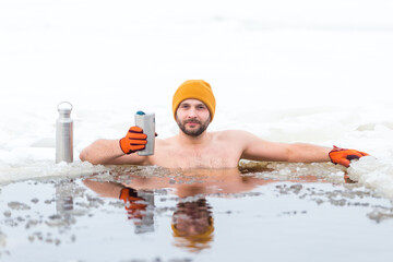 Happy man doing cold baths in winter with hot tea in a thermos. Cold temperatures for ice swimming. Man in orange hat