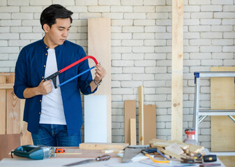 Good looking Asian carpenter working for DIY jobs in carpenter room with several kinds of woods and types of equipment. He pose ahd holding saw.