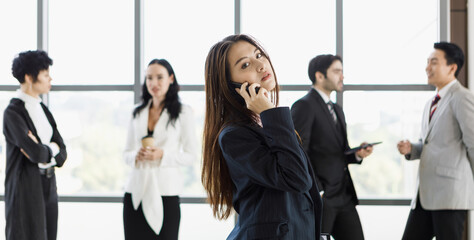 Cute Asian businesswoman using and talking on phone in front of diverse group of businesspeople.