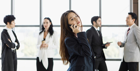 Obraz premium Cute Asian businesswoman using and talking on phone in front of diverse group of businesspeople.