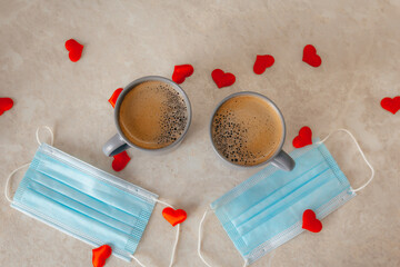 two gray cups of coffee are on the table, along with medical masks and red confetti hearts scattered around. The concept of St. Valentine's Day in a pandemic