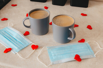 two gray cups of coffee are on the table, along with medical masks and red confetti hearts scattered around. The concept of St. Valentine's Day in a pandemic