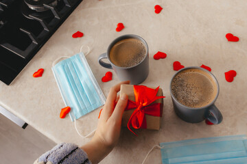 two gray coffee cups stand on the table, a hand holds a gift box with a red bow, medical masks and scattered hearts of red confetti. The concept of St. Valentine's Day in a pandemic