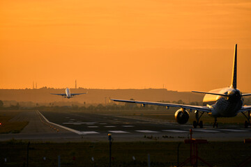 Aeropuerto con dos aviones a la vez sobre al pista de aterrizaje, uno despegando y otro esperando en un horizonte anaranjado.