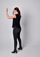 Full length portrait of woman with red hair in a ponytail, wearing black t-shirt and denim pants. Standing pose, hands reaching out with back to the camera the camera, against a  studio background.
