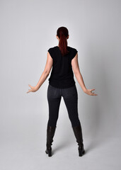 Full length portrait of woman with red hair in a ponytail, wearing black t-shirt and denim pants. Standing pose, hands reaching out with back to the camera the camera, against a  studio background.
