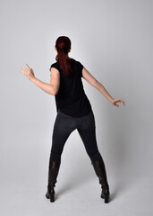 Full length portrait of woman with red hair in a ponytail, wearing black t-shirt and denim pants. Standing pose, hands reaching out with back to the camera the camera, against a  studio background.