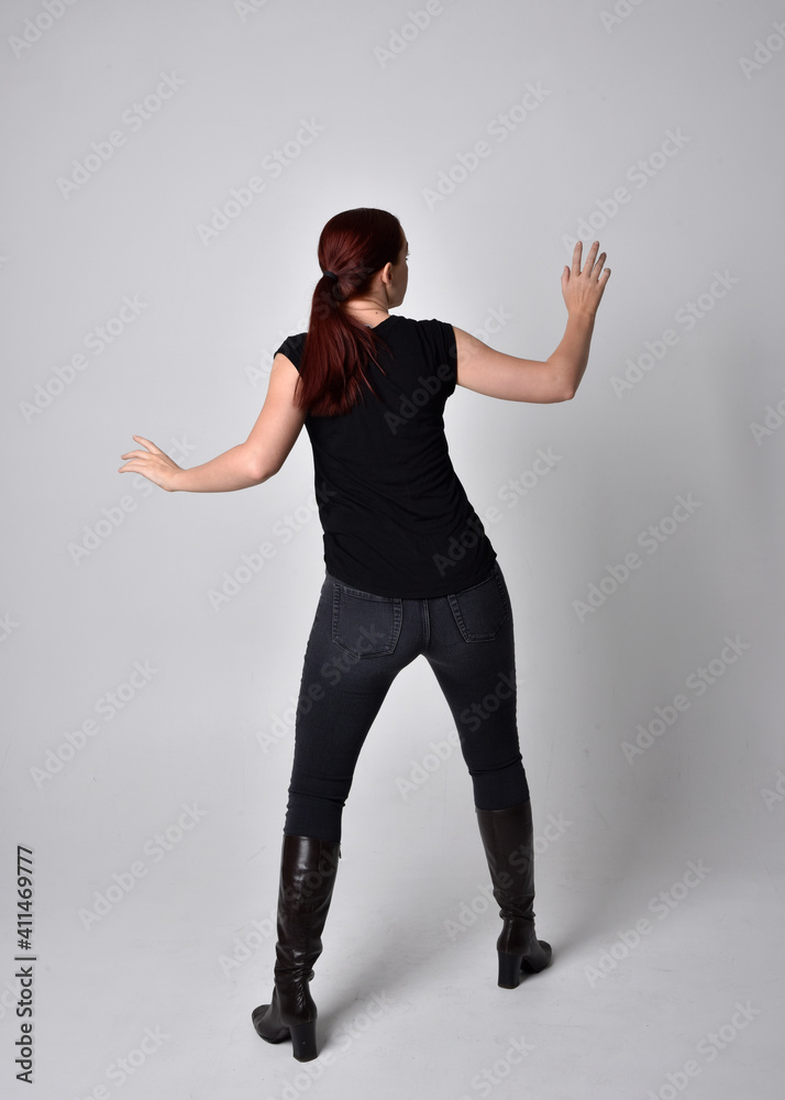 Sticker Full length portrait of woman with red hair in a ponytail, wearing black t-shirt and denim pants. Standing pose, hands reaching out with back to the camera the camera, against a  studio background.