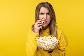 Photo of attractive lady holds plate of popcorn, eats it with disgruntled face. Wears casual yellow hoody, isolated yellow color background