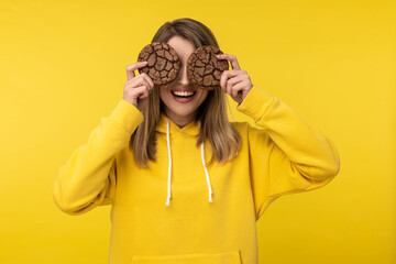 Photo of attractive lady holds cookies and covers eyes up. Wears casual yellow hoody, isolated yellow color background