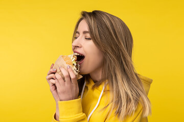 Photo of attractive lady poses in a surprised way bites burger. Wears casual yellow hoody, isolated yellow color background