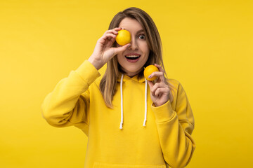 Fototapeta na wymiar Photo of attractive lady poses in a cute way with lemons, cover one eye up with. Wears casual yellow hoody, isolated yellow color background