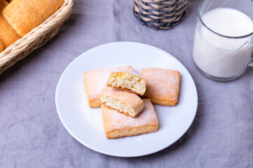Cookies with powdered sugar on a white plate. In the background a basket of cookies and a cup of milk. Close-up.
