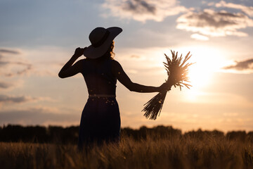 Silhouette of a girl with an armful of spikelets of wheat in her hands against the backdrop of the setting sun