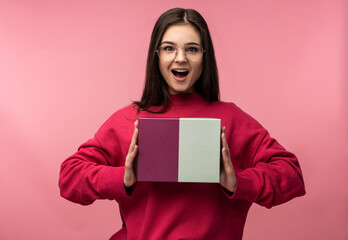 Photo of attractive lady in glasses holds box with present, surprised and gapes, wear casual pink sweater white pants isolated pink color background.