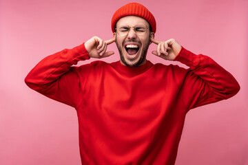Naklejka premium Photo of attractive man with beard in glasses and red clothing. Male screams and plugs ears, isolated over pink background