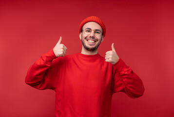 Naklejka premium Photo of attractive man with beard in glasses and red clothing. Male gives thumb up and smiles, isolated over red background