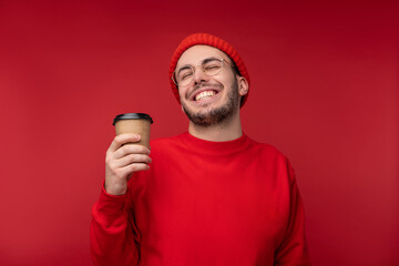 Photo of attractive man with beard in glasses and red clothing. Happy man holds cup of coffee and looks cute, isolated over red background