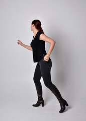 Simple full length portrait of woman with red hair in a ponytail, wearing casual black tshirt and jeans. Standing pose with back to the camera the camera, against a  studio background.