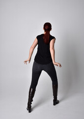 Simple full length portrait of woman with red hair in a ponytail, wearing casual black tshirt and jeans. Standing pose with back to the camera the camera, against a  studio background.