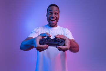 Excited african american guy playing video games with joystick in neon lighting