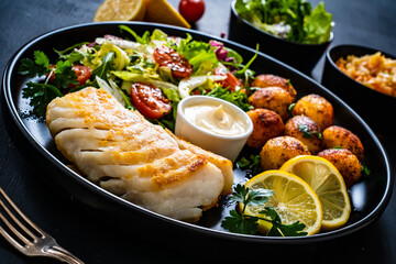 Fish dish - fried cod fillet with potatoes and vegetable salad on black wooden table
