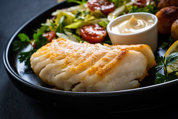 Fish dish - fried cod fillet with potatoes and vegetable salad on black wooden table
