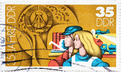  a postage stamp from Germany, GDR showing a portrait of a man and woman in work clothes in front of agricultural facilities and buildings with the GDR emblem. 35 years of the GDR