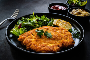 Breaded fried pork chop and fresh vegetables on black table

