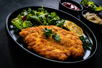 Breaded fried pork chop and fresh vegetables on black table


