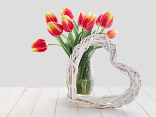 Bunch of red yellow stripe tulips in glass vase. Whitewashed wattle rattan wreath in shape of heart. Spring decorations on off white, natural rustic wooden table.