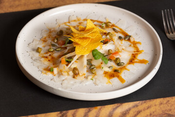 White plate with Cod Carpaccio with chili vinaigrette, pumpkin seeds and chips.