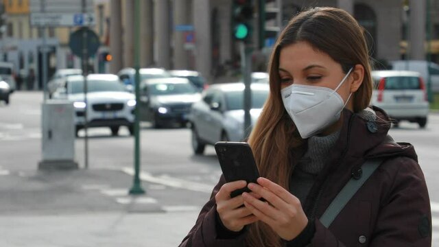 Portrait of young woman using smartphone wearing FFP2 KN95 protective mask and winter jacket outdoors