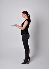 Simple full length portrait of woman with red hair in a ponytail, wearing casual black tshirt and jeans. Standing pose with in side profile, against a  studio background.