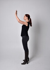 Simple full length portrait of woman with red hair in a ponytail, wearing casual black tshirt and jeans. Standing pose with in side profile, against a  studio background.