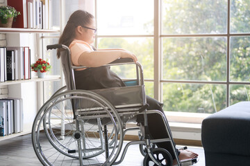 A lonely child Asian autistic girl sits in a wheelchair and looks out the window to feel lonely at home during the COVID-19 epidemic.