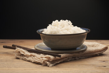 Cooked Thai jasmine rice in a ceramic bowl with chopsticks placed on an old wooden table against a black background.
