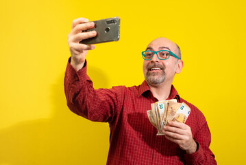 Middle-aged Caucasian man holding a bunch of 50 euro bills in one hand and taking a selfie with the...