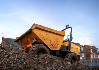 Mini yellow dumper with roll bar and orange flashing lights and tipper front bucket  parked on construction mound of soil on new houses industrial building site 