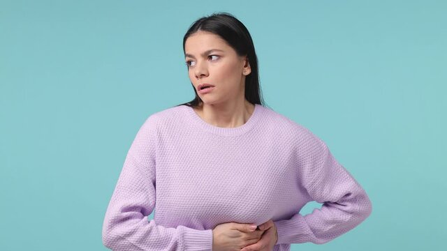 Sick ill brunette young latin woman 20s in violet sweater posing isolated on blue background studio. People lifestyle concept. Put hands on abdomen with stomach-ache griping bellyache feel bad seedy