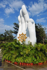 White sculpture in the form of a hand on a background of green trees and blue sky