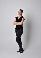 Simple full length portrait of woman with red hair in a ponytail, wearing casual black tshirt and jeans. Standing pose facing front on, against a  studio background.