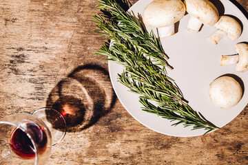 Flat lay view of glass with red wine and white plate with the minimalist vegan food - champignon mushrooms and rosemary branches against old wooden backdrop with texture. Concept idea of healthy life.
