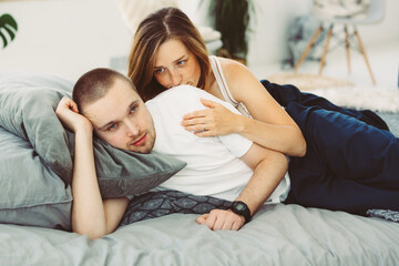 young married couple in clothes lie on the bed with gray sheets. A woman hugs a man from behind.