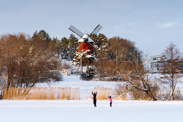 People ice skating on a frozen lake in front of a red, snowy windmill, Stockholm, Sweden