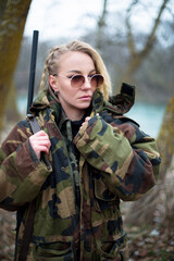 caucasian woman in camouflage suit and sunglasses with shotgun on the shoulder near the river