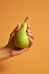 Studio shot of female hand holding ripe green pear isolated over orange background. Healthy eating and diet concept