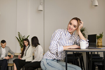 young blonde woman drinks coffee in the office and a group of her Colleagues sit at the table on blurred background