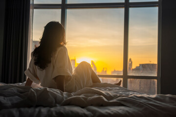 The sun shines through window in the morning. Beautiful young woman sitting on bed. Happy young...