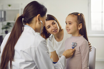 Young woman doctor pediatrician in medical uniform examining little smiling girl with stethoscope with mother sitting near during checkup and consultation in medical clinic office. Medicare for kids